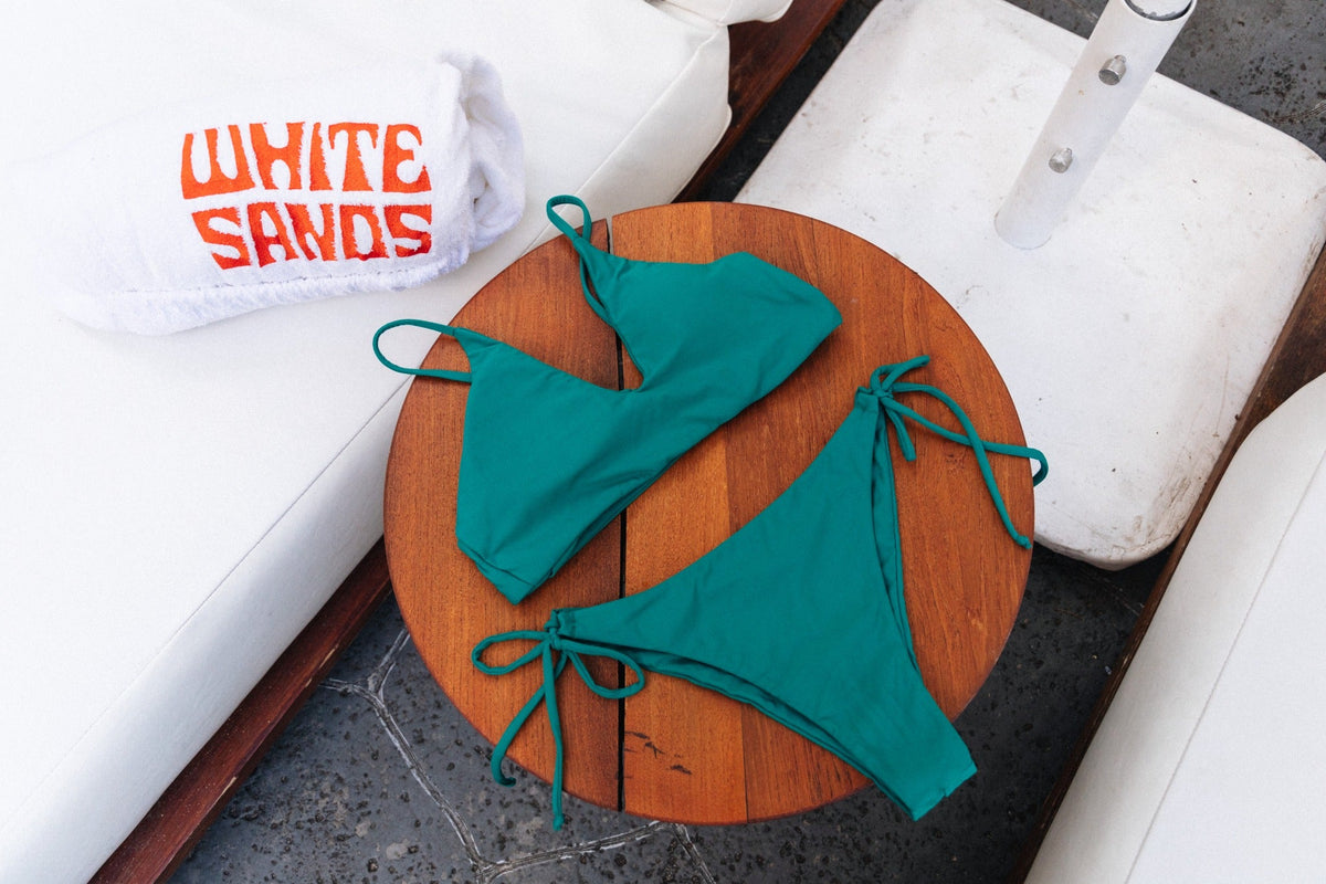 Teal triangle bikini arranged on an orange board beside a White Sands cap.